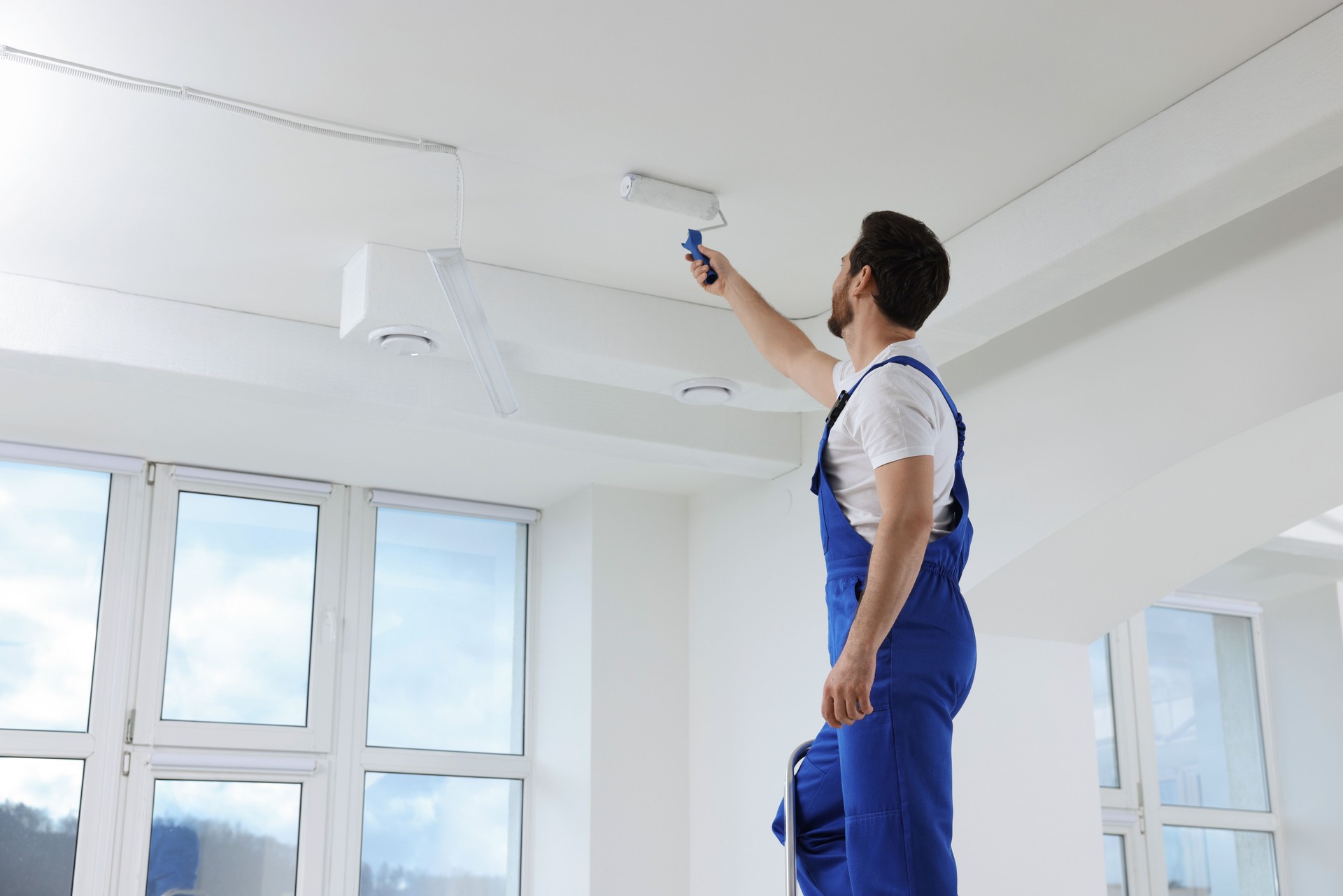 Handyman with roller painting ceiling on step ladder in room